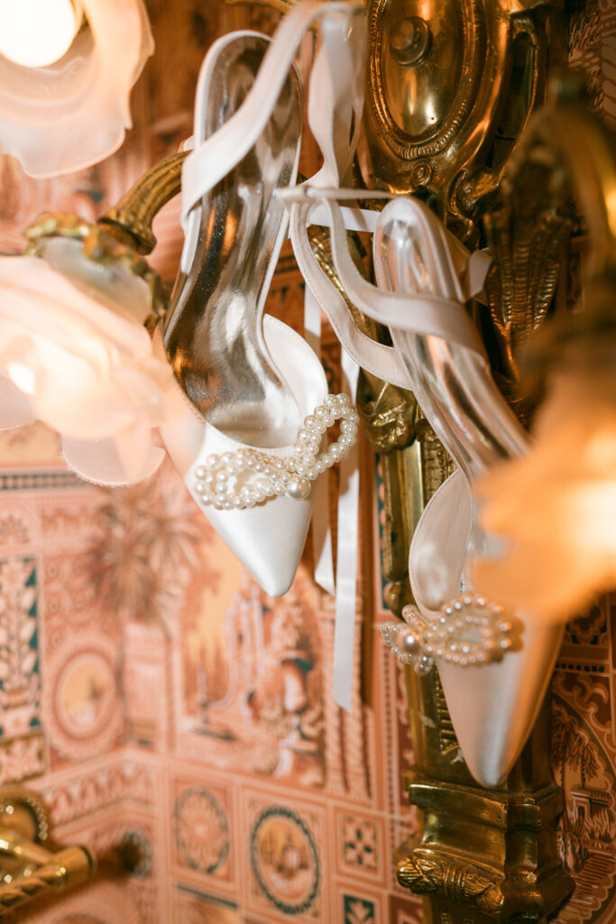wedding detail shot of bride's shoes hanging on vintage flower light fixture with pink wallpaper