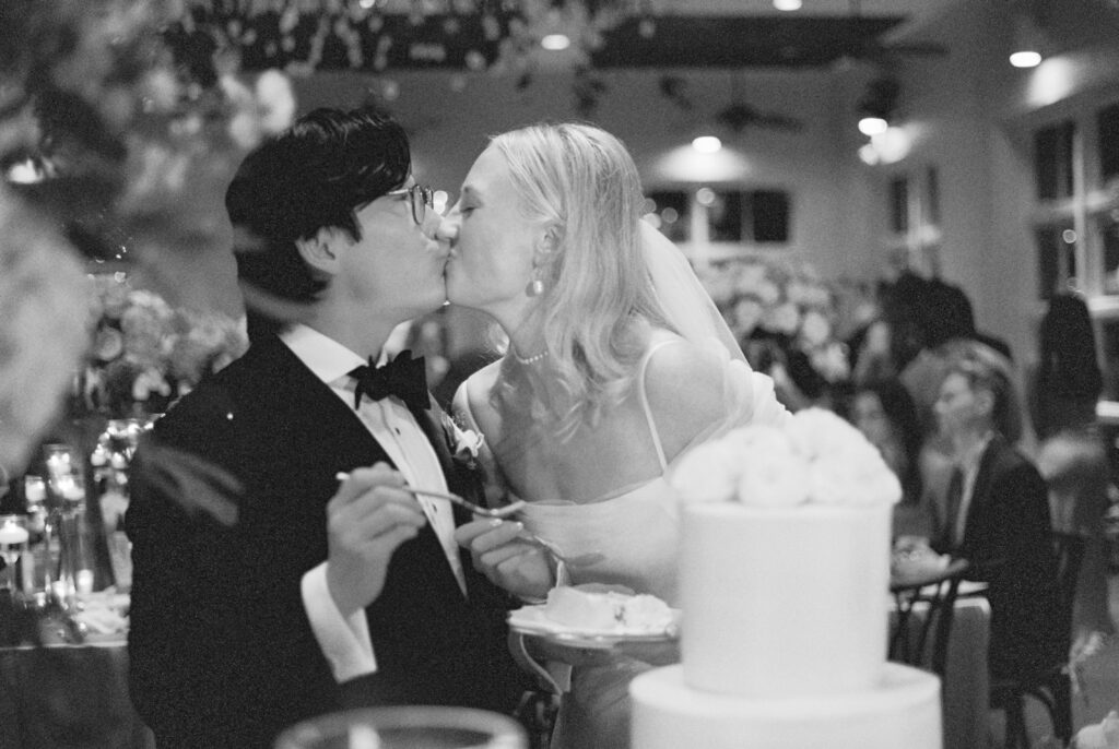 bride and groom couple kissing over slice of wedding cake in crowded reception room on film wedding photography